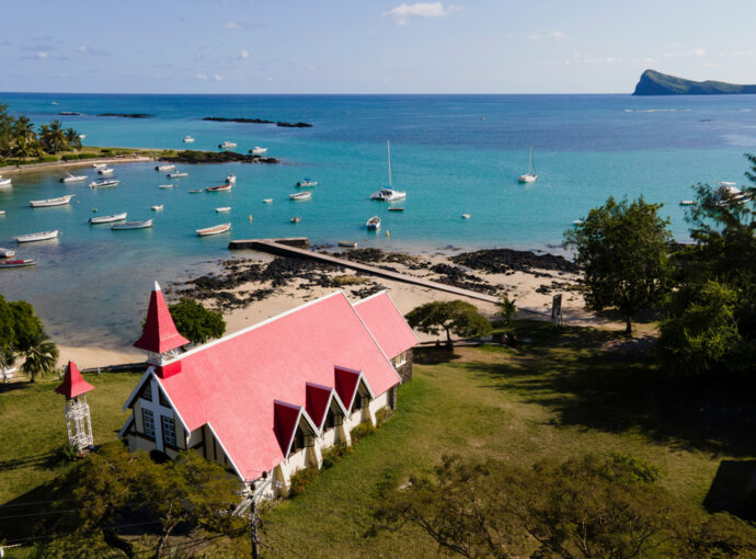 Aerial view of a seaside village with a white church and bright red roof, turquoise bay, and numerous moored boats near the beach.
