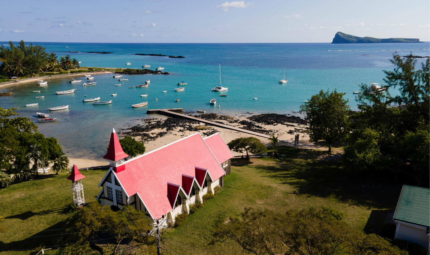 Aerial view of a seaside village with a white church and bright red roof, turquoise bay, and numerous moored boats near the beach.